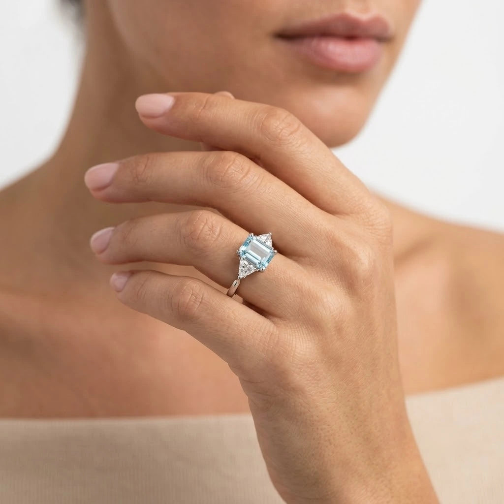 FineDiam emerald cut blue gemstone ring with sparkling triangular side stones and a polished white metal band.