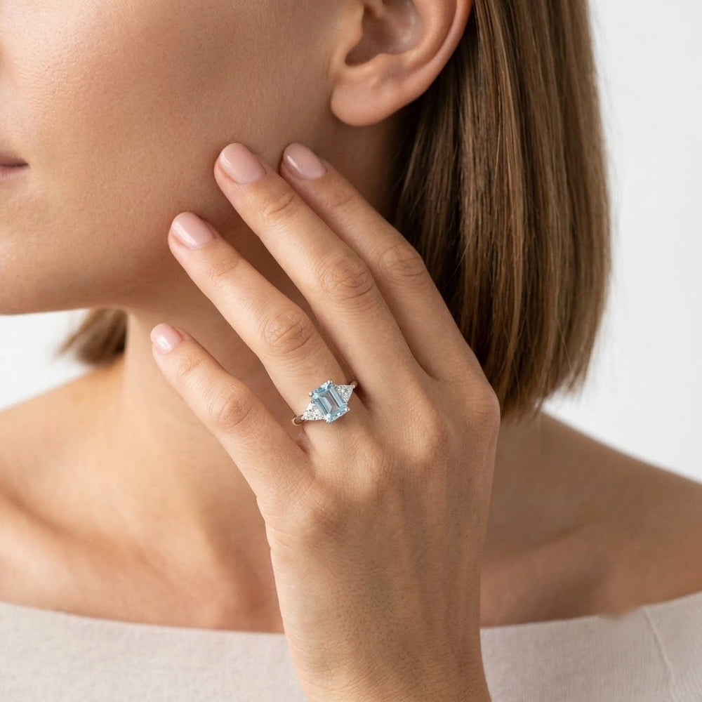  FineDiam blue emerald cut gemstone ring featuring two triangular side stones in a white metal setting.
