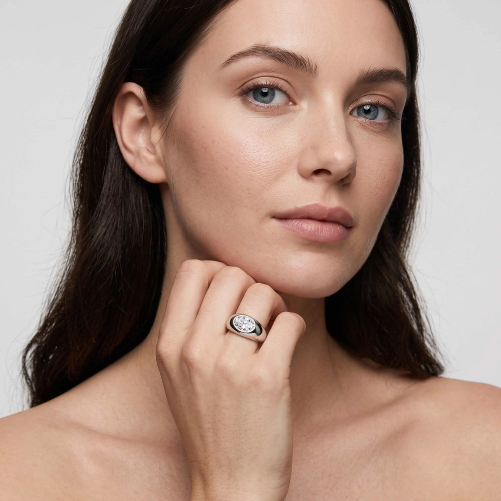 A close-up portrait of a woman wearing a silver-toned ring with a round clear gemstone on her finger, set against a soft light gray background.

