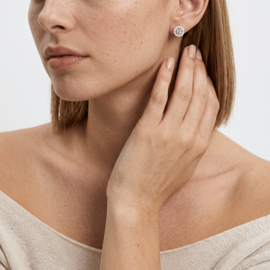 Close-up view of a woman with her hand resting near her jaw, showing a round silver halo stud earring with clear crystals.