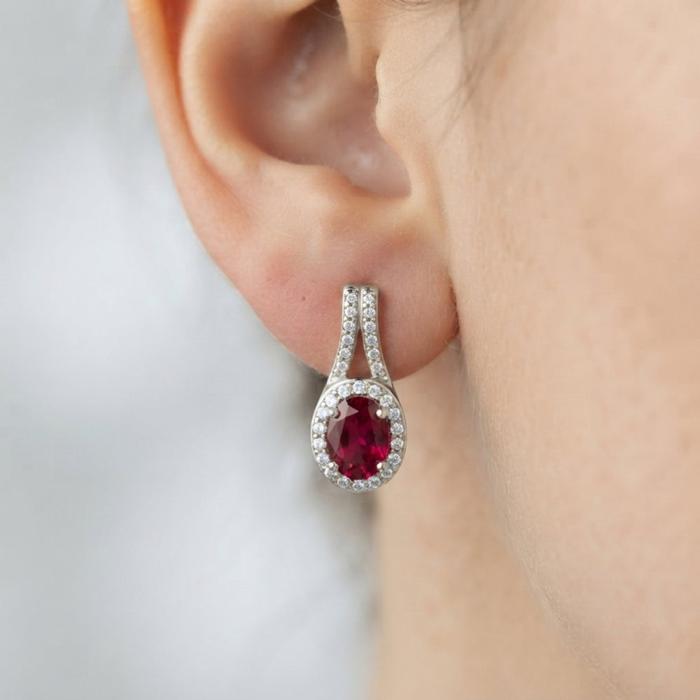 Extreme close-up of a woman's ear wearing a white gold drop earring featuring an oval red ruby stone surrounded by diamond halos.